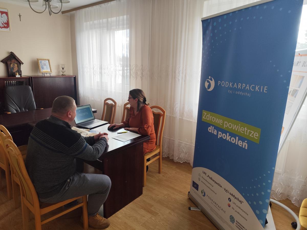 Dwie osoby siedzą przy biurku. Na pierwszym planie niebieski baner z napisem: "Podkarpackie: żyj i oddychaj. Zdrowe powietrze dla pokoleń".